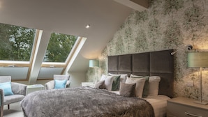 A bedroom with a large bed, two armchairs, a nightstand, a lamp, and a view of trees through skylights. - Orsett Hall Hotel (Grays)