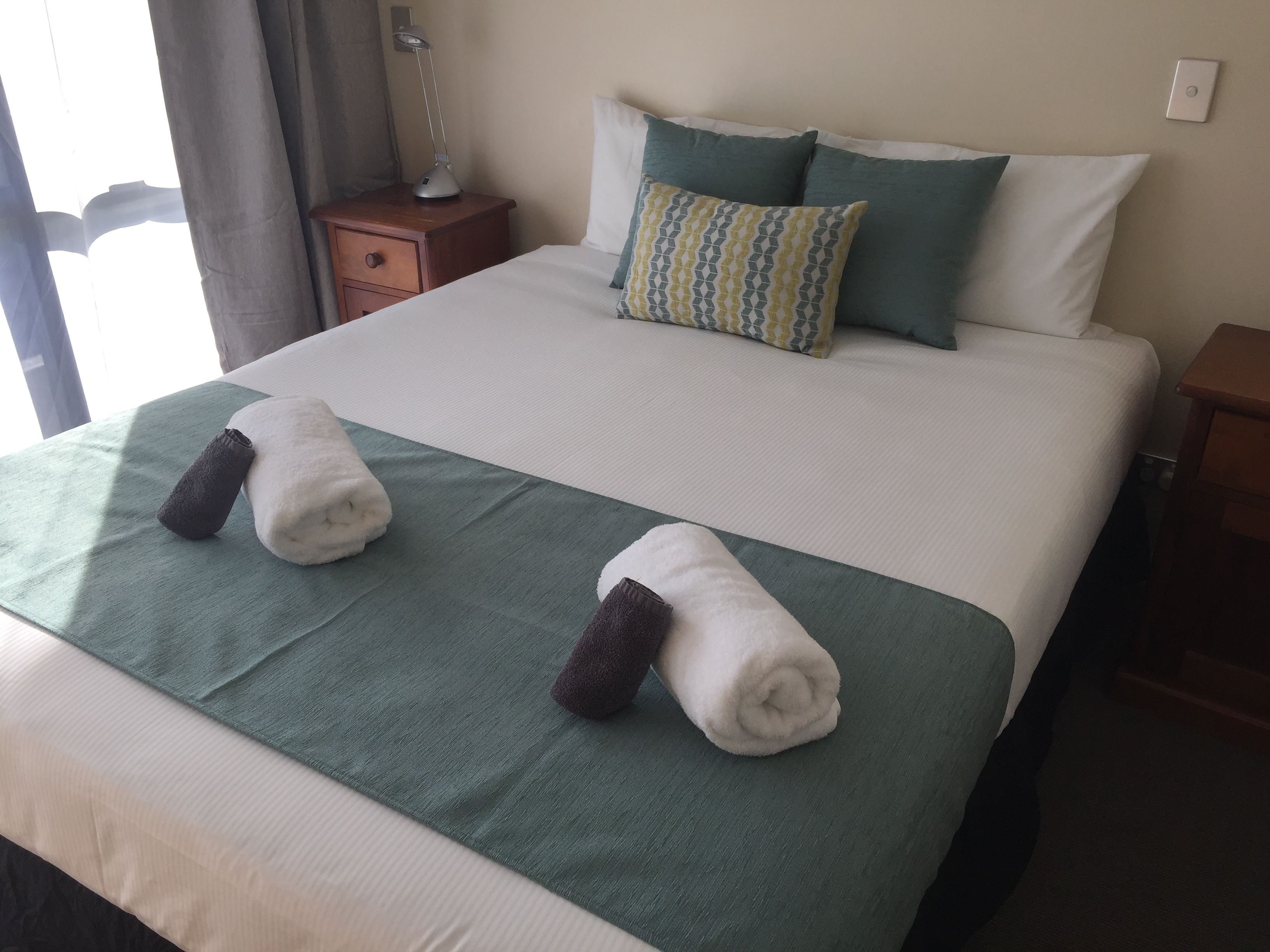 A neatly made bed with a green bedspread, two rolled towels, and a patterned pillow.