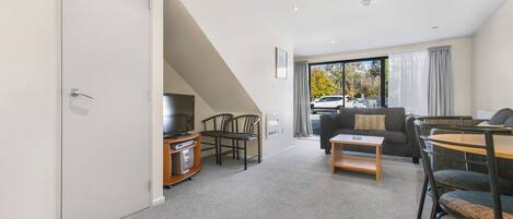 A modern living room with a sofa, coffee table, and chairs. There is a TV on a stand and a window with curtains.