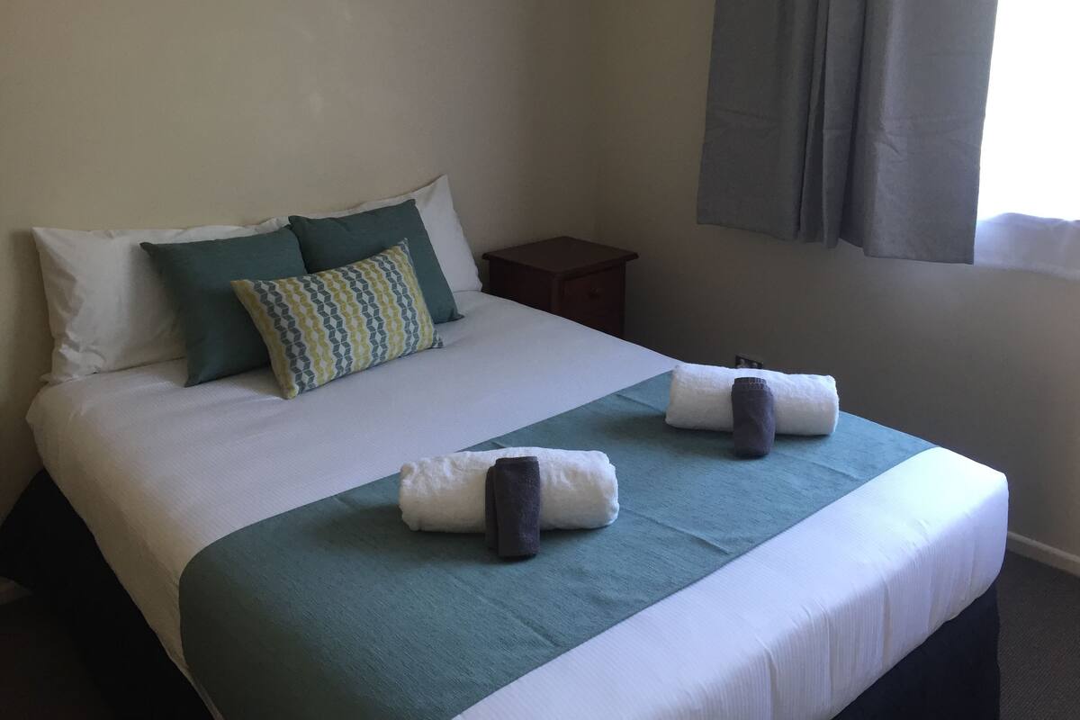A neatly made bed with a green and white bedspread, two pillows, and two rolled towels.