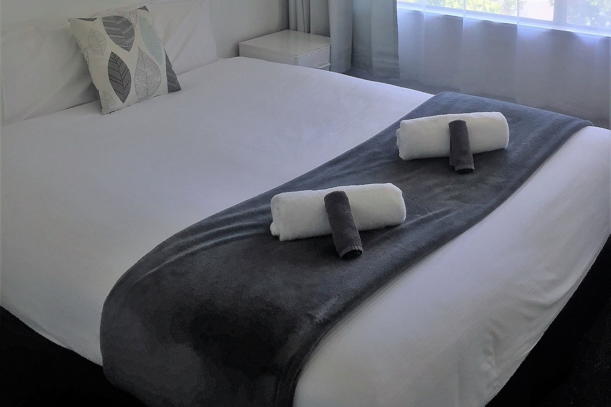 A neatly made bed with a grey blanket and two rolled towels, a bedside table, a pillow with a leaf pattern, and a window with curtains.