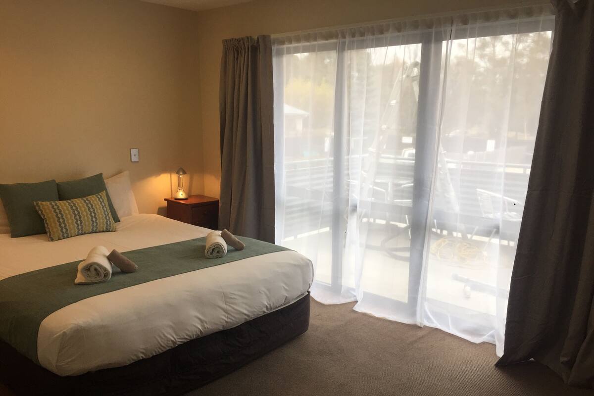 A hotel room with a large bed, a nightstand with a lamp, and a view of the outdoor area through sheer curtains.
