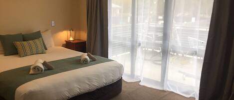 Habitación de hotel con una cama grande, una mesita de noche con lámpara y vistas al exterior a través de cortinas translúcidas.