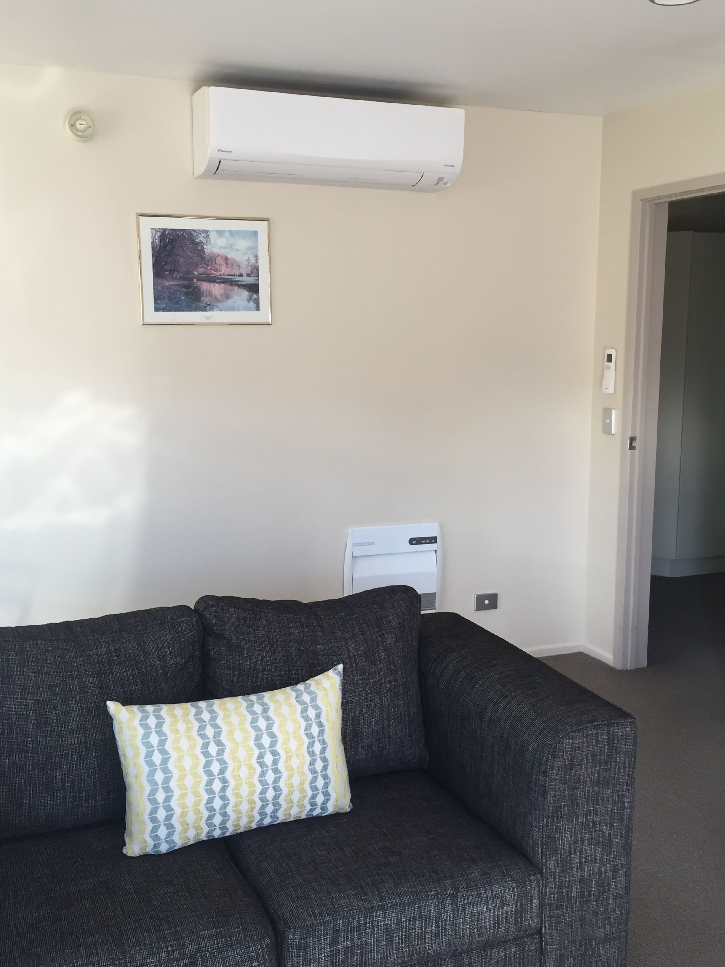 A living room with a dark grey sofa, a patterned cushion, an air conditioning unit, a framed picture on the wall, and a door leading to another room.