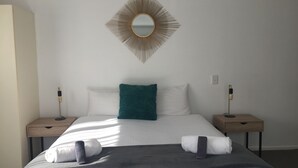 A neatly made bed with white and teal pillows, a dark throw blanket, and two towels. A gold-framed mirror hangs above the bed. - Fairway Motel and Apartments (Wanaka)