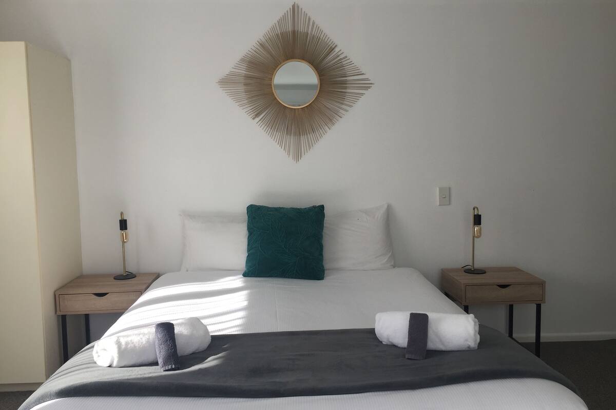 A neatly made bed with white and teal pillows, a dark throw blanket, and two towels. A gold-framed mirror hangs above the bed.