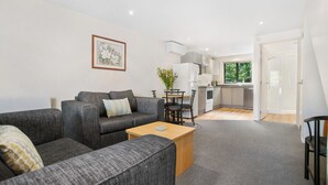 A modern living room with grey sofas, a wooden coffee table, and a dining area with a small table and chairs. A kitchen is visible in the background with white cabinets and a window. - Fairway Motel and Apartments (Wanaka)