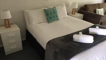 Een net opgemaakt bed met witte lakens, een kussen met een groen patroon en opgerolde handdoeken op een donkere spreidek. Twee nachtkastjes met lampen, een bruine fauteuil en een witte ladekast.