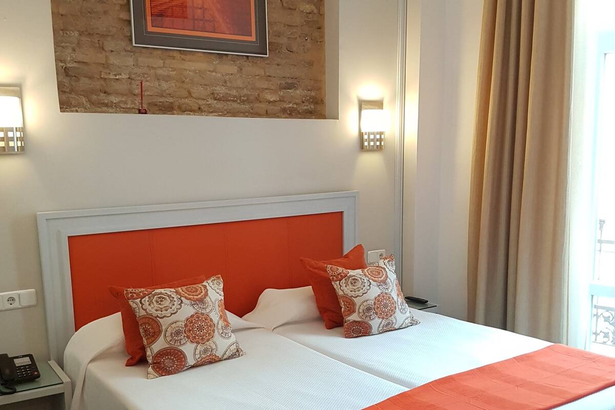 A double bed with an orange headboard and white bedding, two patterned pillows, a red throw blanket, a nightstand with a phone, and a brick wall with a framed picture above.