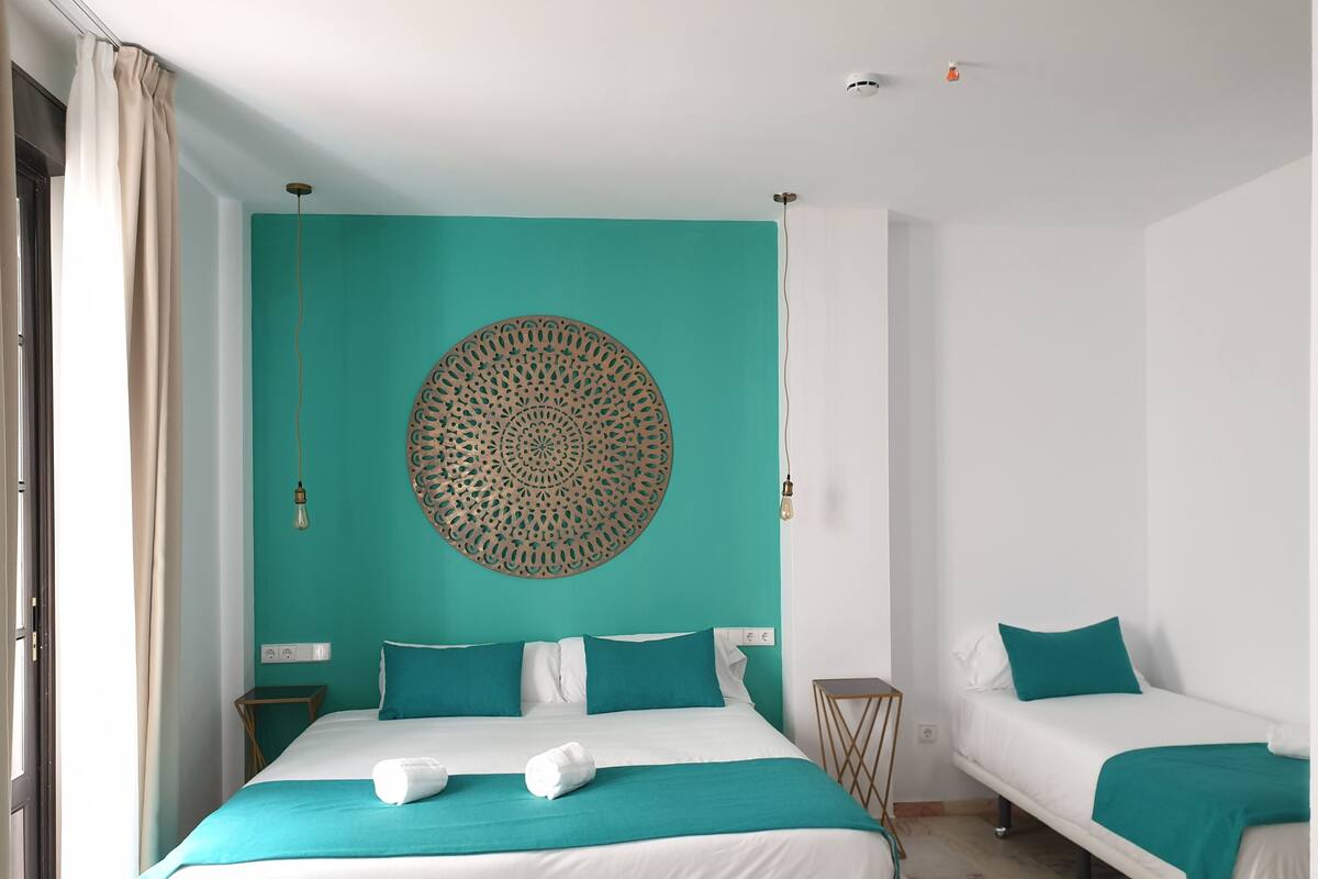 A hotel room with a bed, a smaller bed, teal and white bedding, a decorative wall hanging, and a small side table.