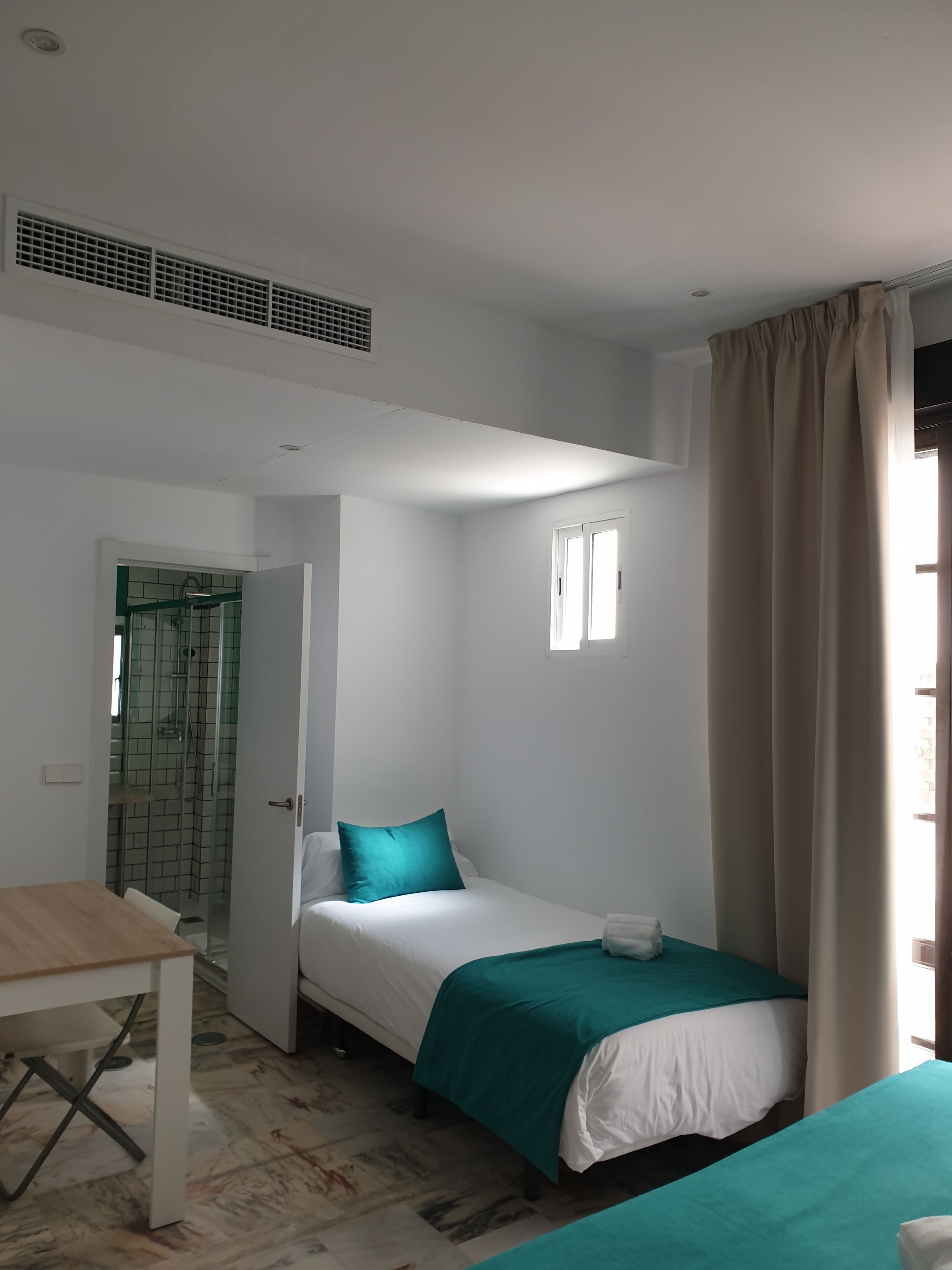 Quarto pequeno e limpo com uma cama de solteiro, uma mesa e um espaço com chuveiro.