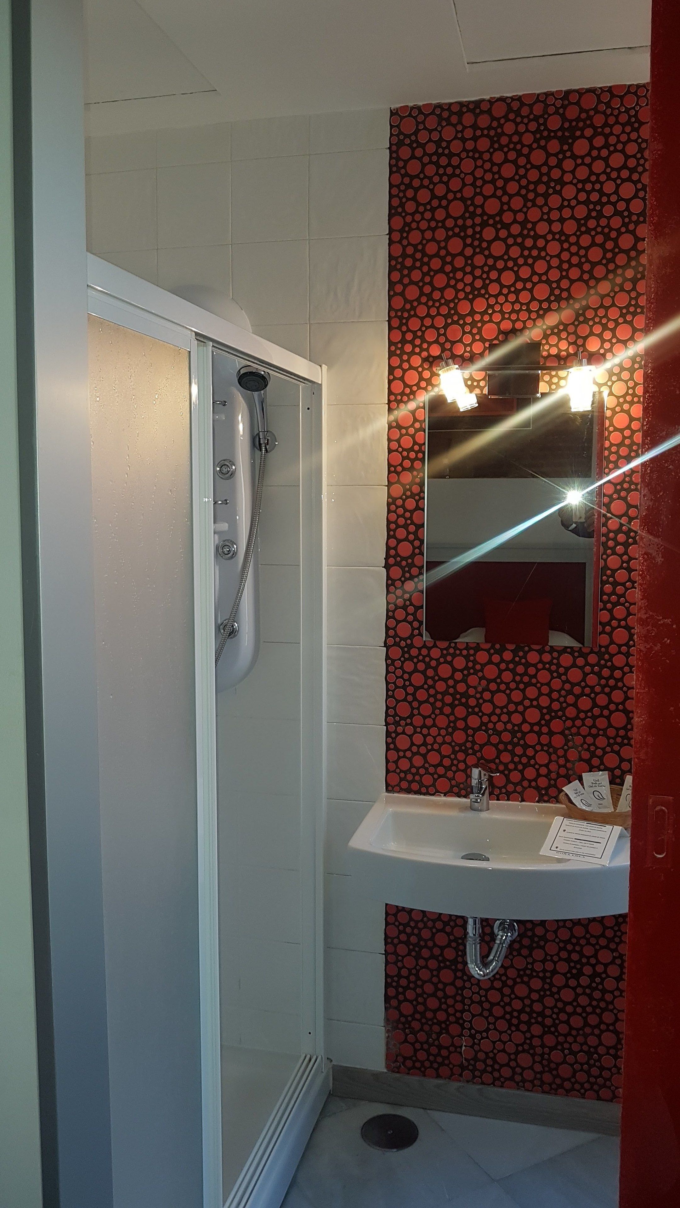 A bathroom with a glass-enclosed shower, a sink with a mirror above it, and a red polka dot wall.
