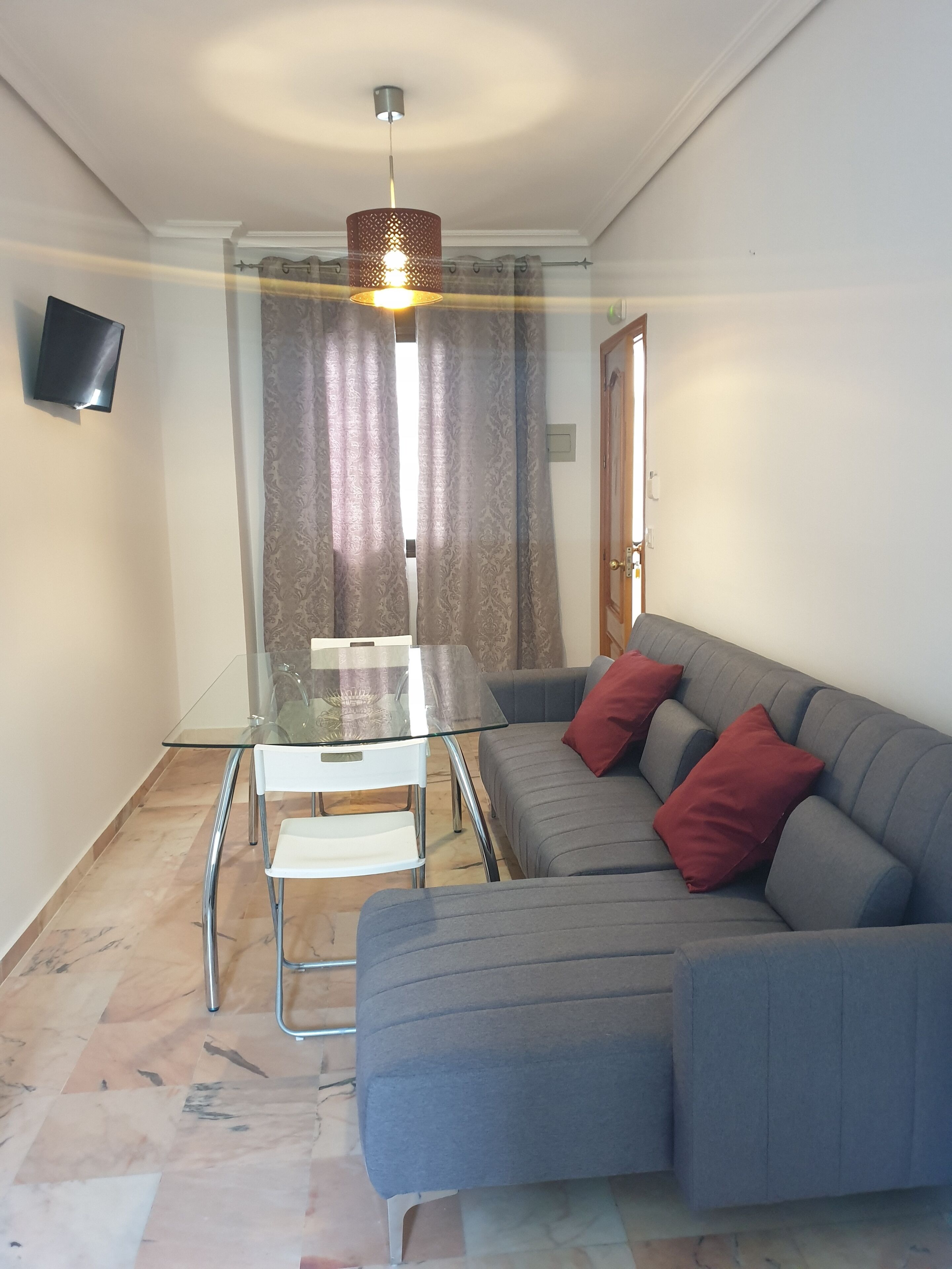 A modern living room with a glass-top dining table, a grey sofa with red cushions, and a pendant light.