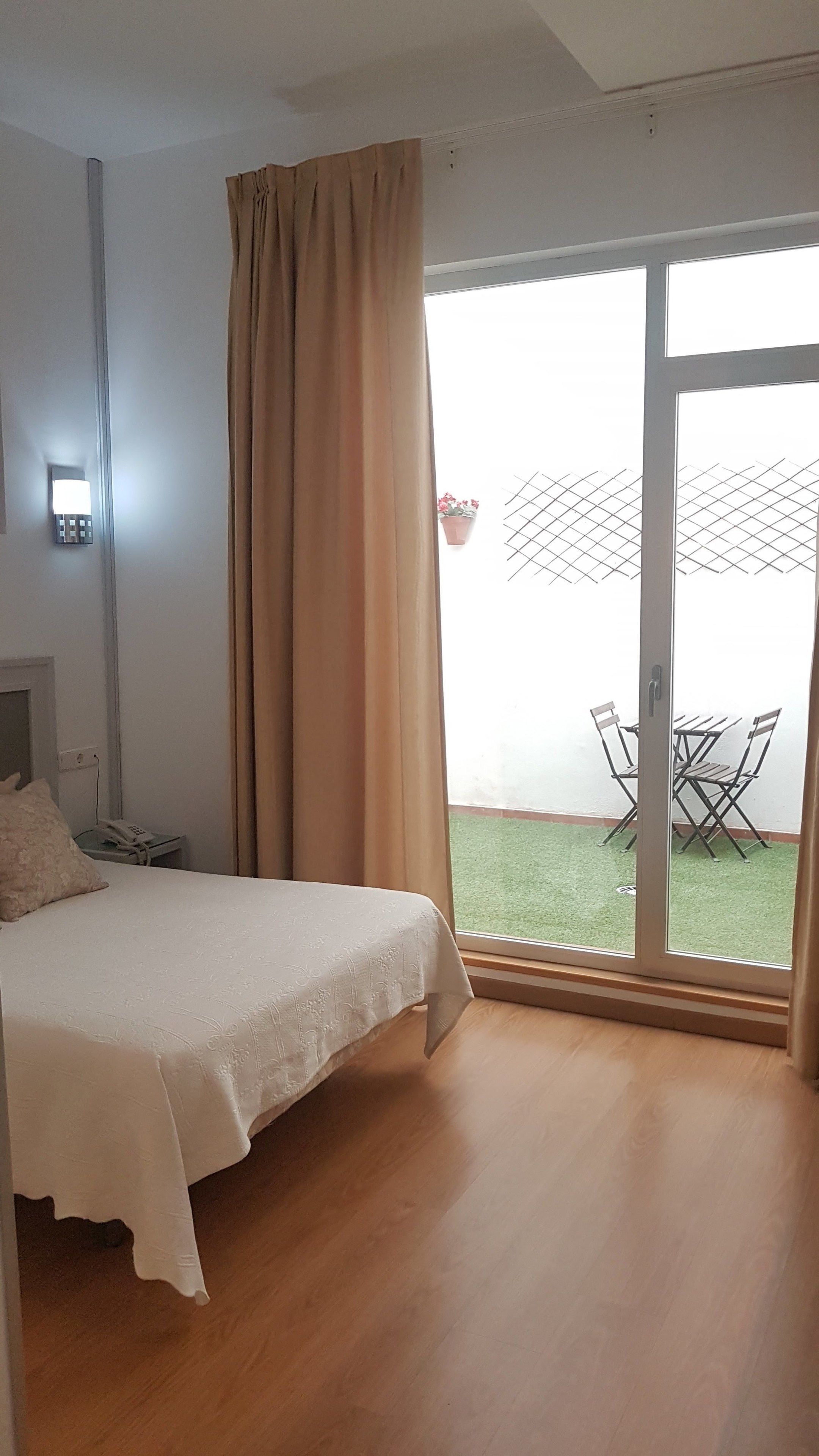 A bedroom with a bed, a bedside phone, a window with curtains, and a view of a patio with a table and chairs.