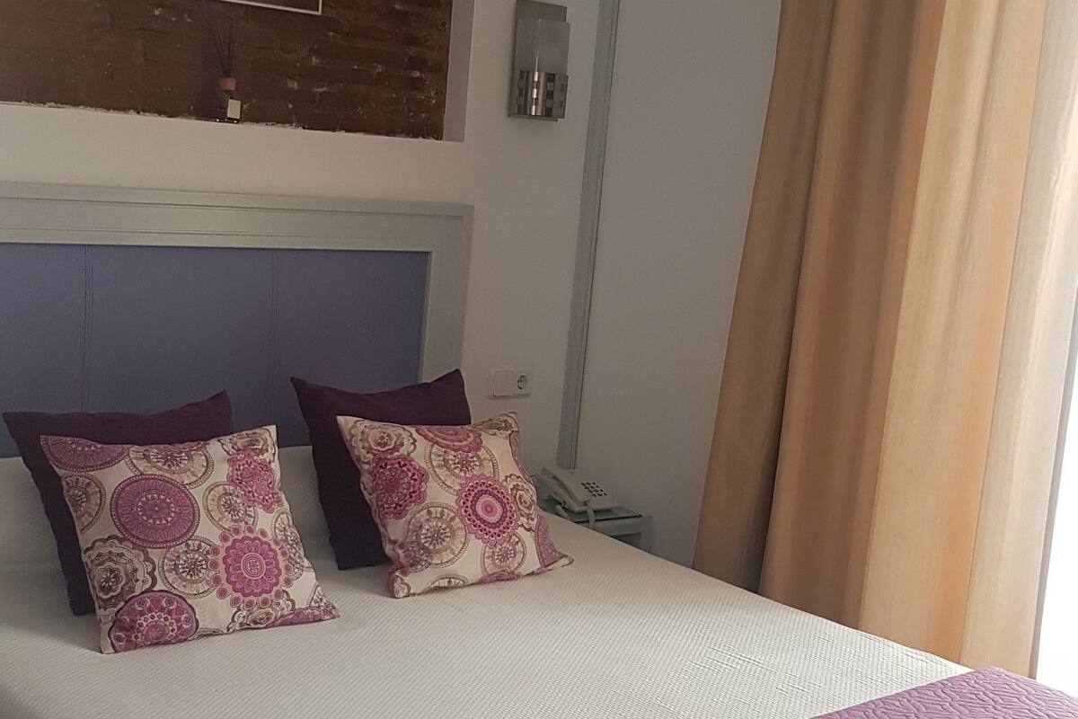 A neatly made bed with a white and purple bedspread, two patterned pillows, and a headboard against a brick wall.