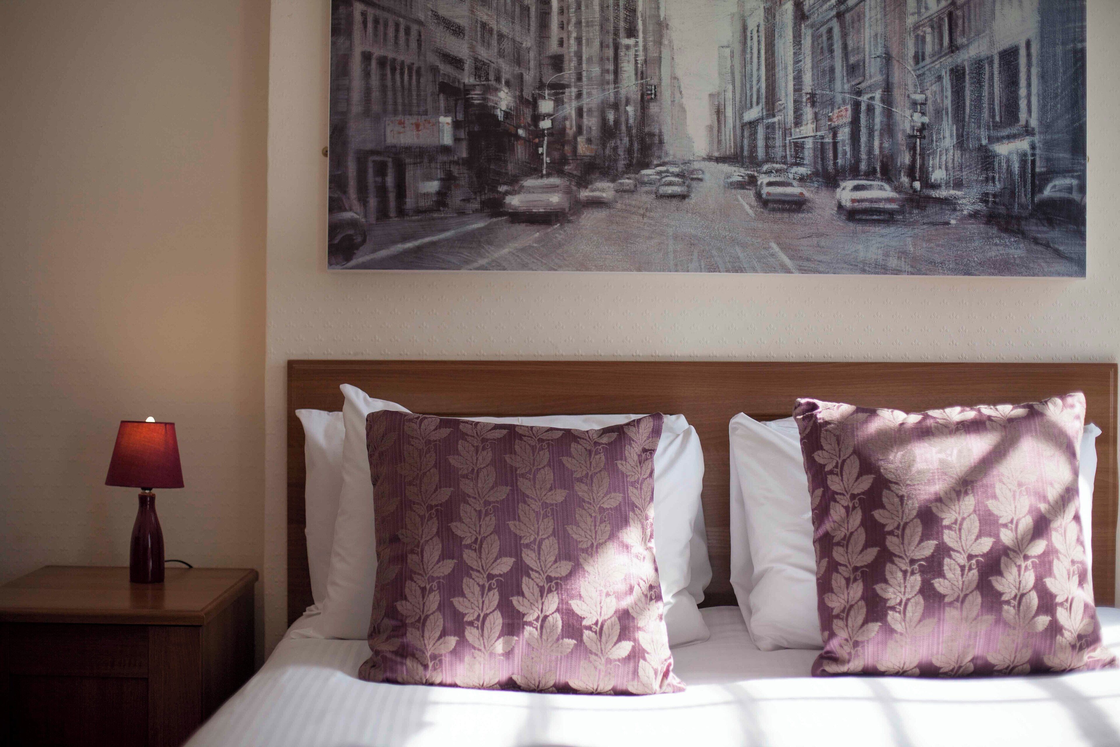 A hotel room with a bed, two pillows, a nightstand with a lamp, and a cityscape painting on the wall.