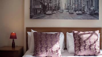A hotel room with a bed, two pillows, a nightstand with a lamp, and a cityscape painting on the wall.