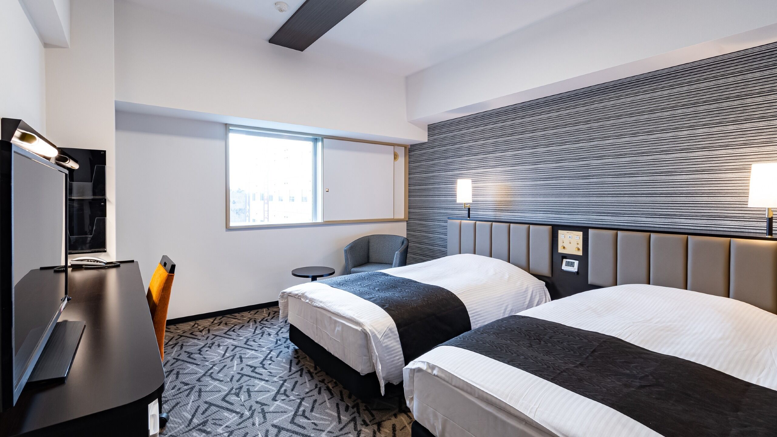 A hotel room with two beds, a desk, a chair, a TV, and a window.