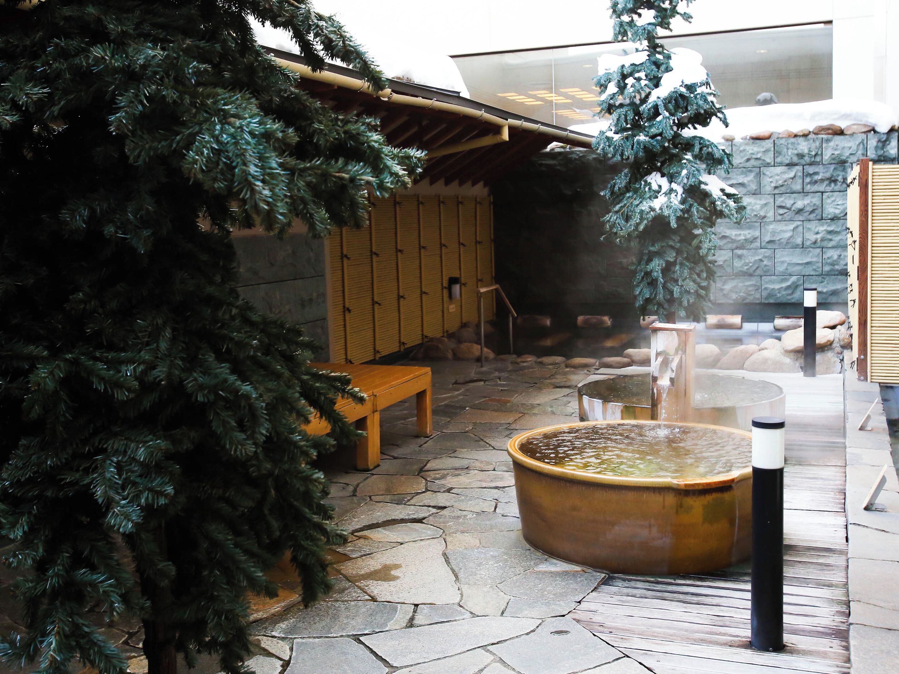 A stone-floored spa area with a wooden bench, a hot spring, and a pine tree.