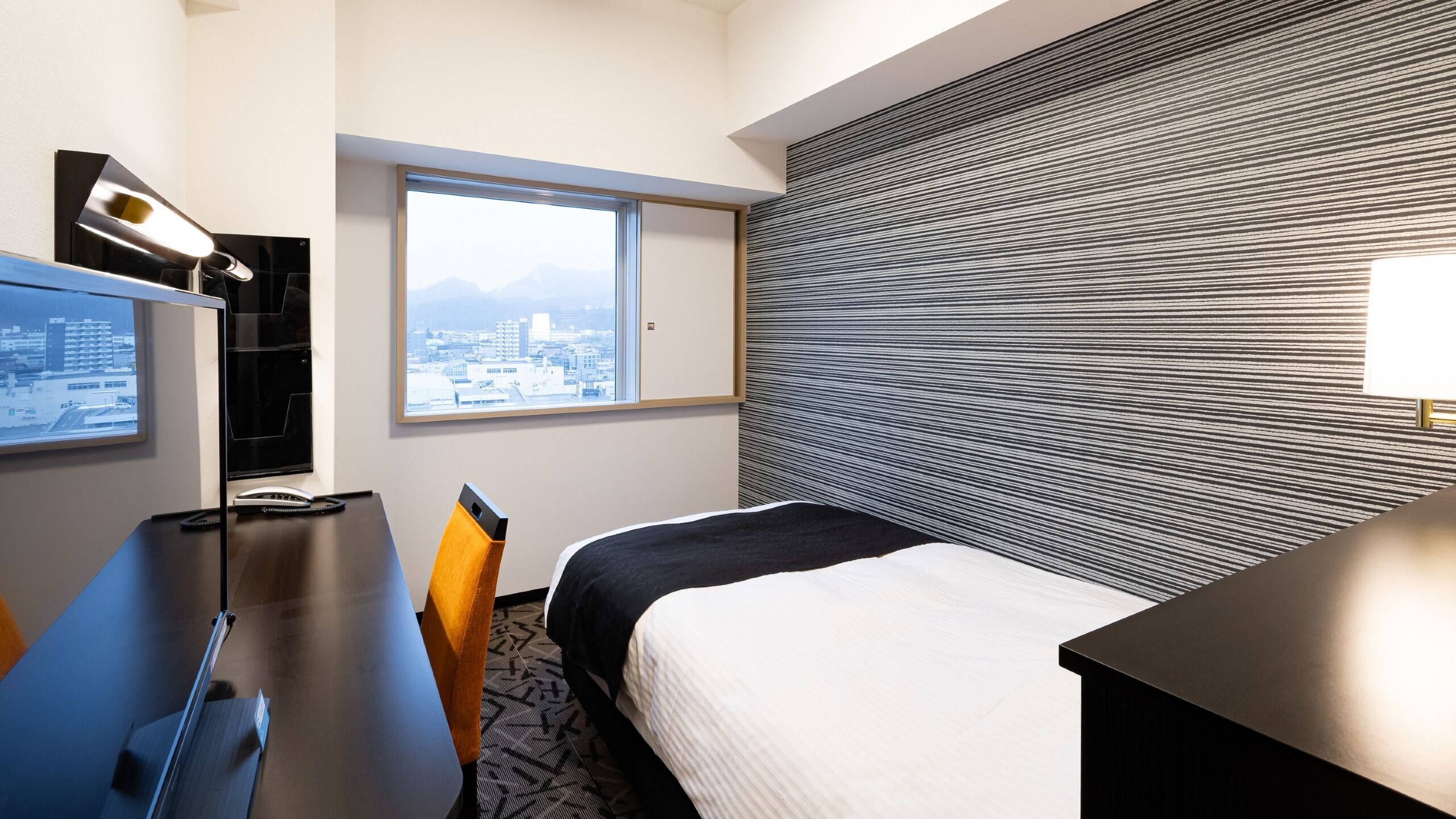 A hotel room with a bed, a desk, a chair, a TV, and a window with a city view.