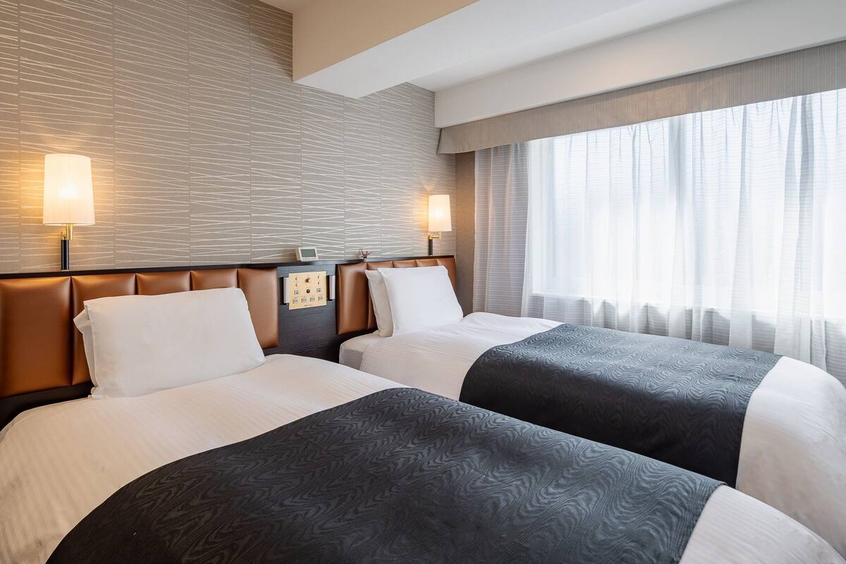 A hotel room with two beds, a control panel, and a window with sheer curtains.