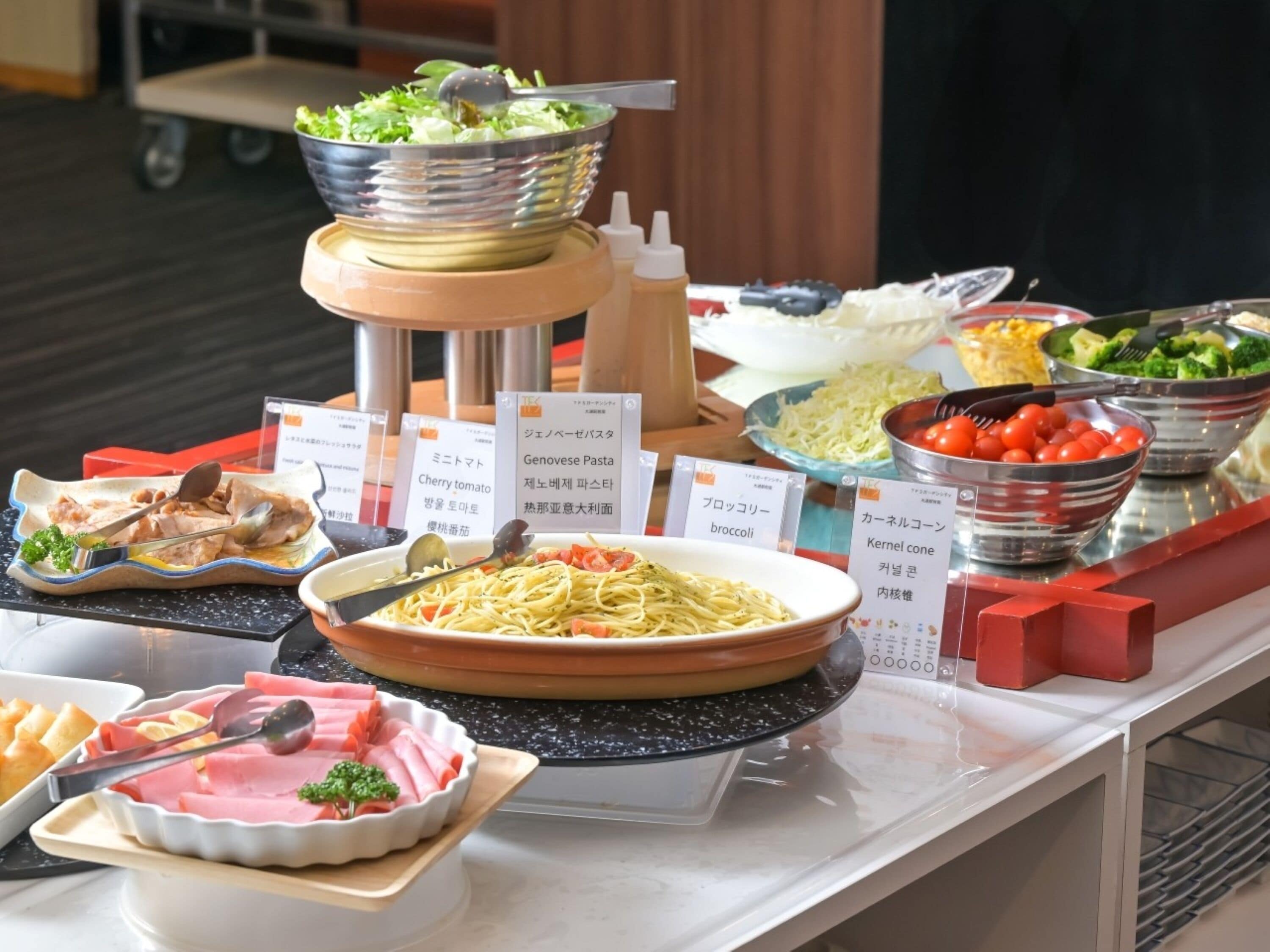 A buffet with various dishes including pasta, vegetables, and meats, with labels in a foreign language.