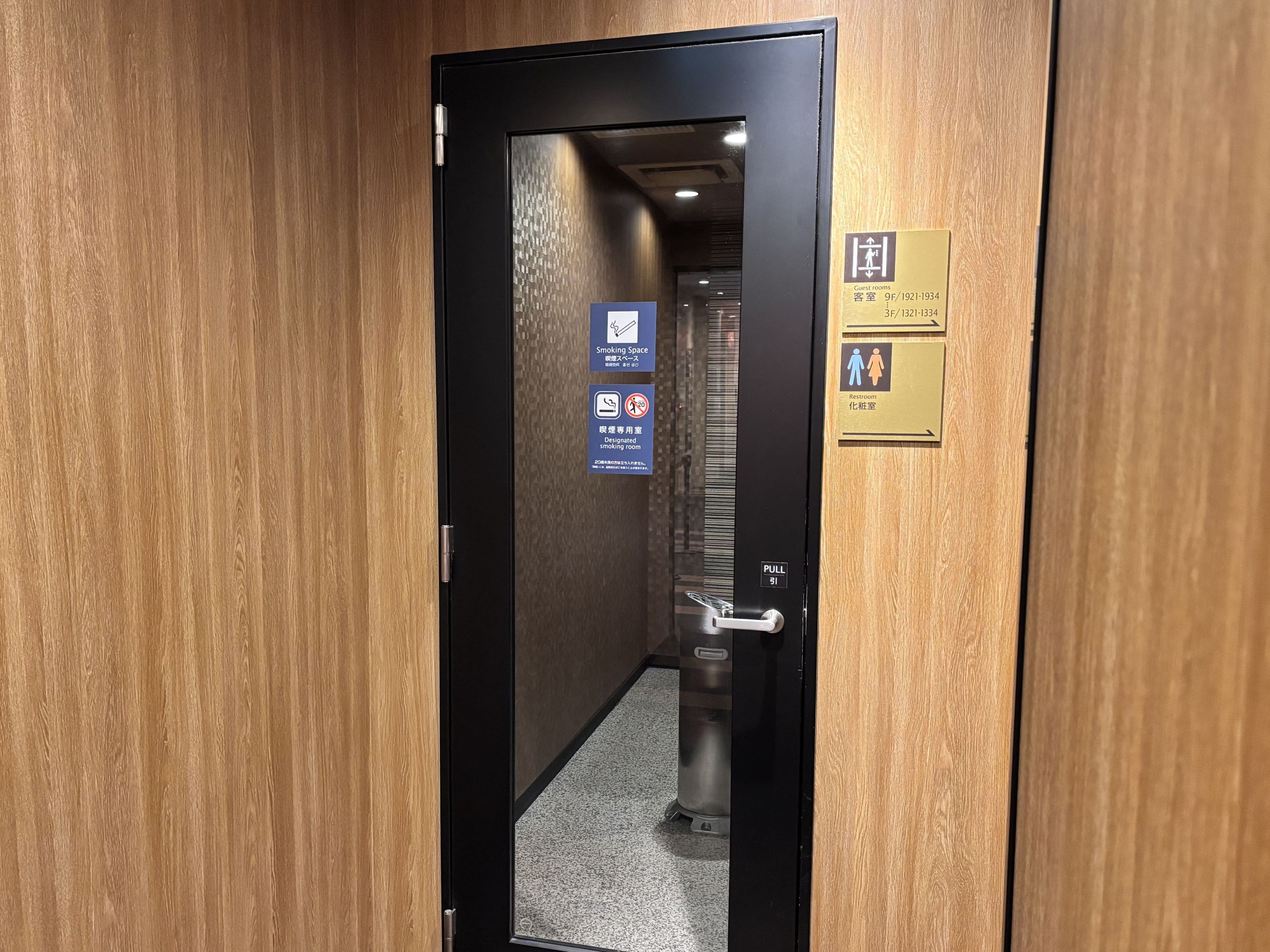 Pasillo de hotel con puerta corredera, un cubo de basura y carteles que indican habitaciones de huéspedes, zona para fumadores y baños.