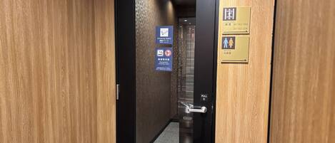 Pasillo de hotel con puerta corredera, un cubo de basura y carteles que indican habitaciones de huéspedes, zona para fumadores y baños.
