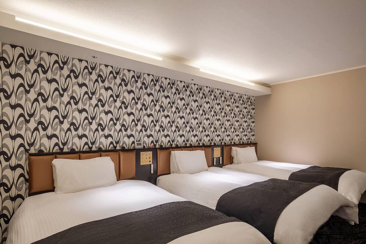 A hotel room with three beds, a patterned wallpaper, a control panel, and a carpeted floor.