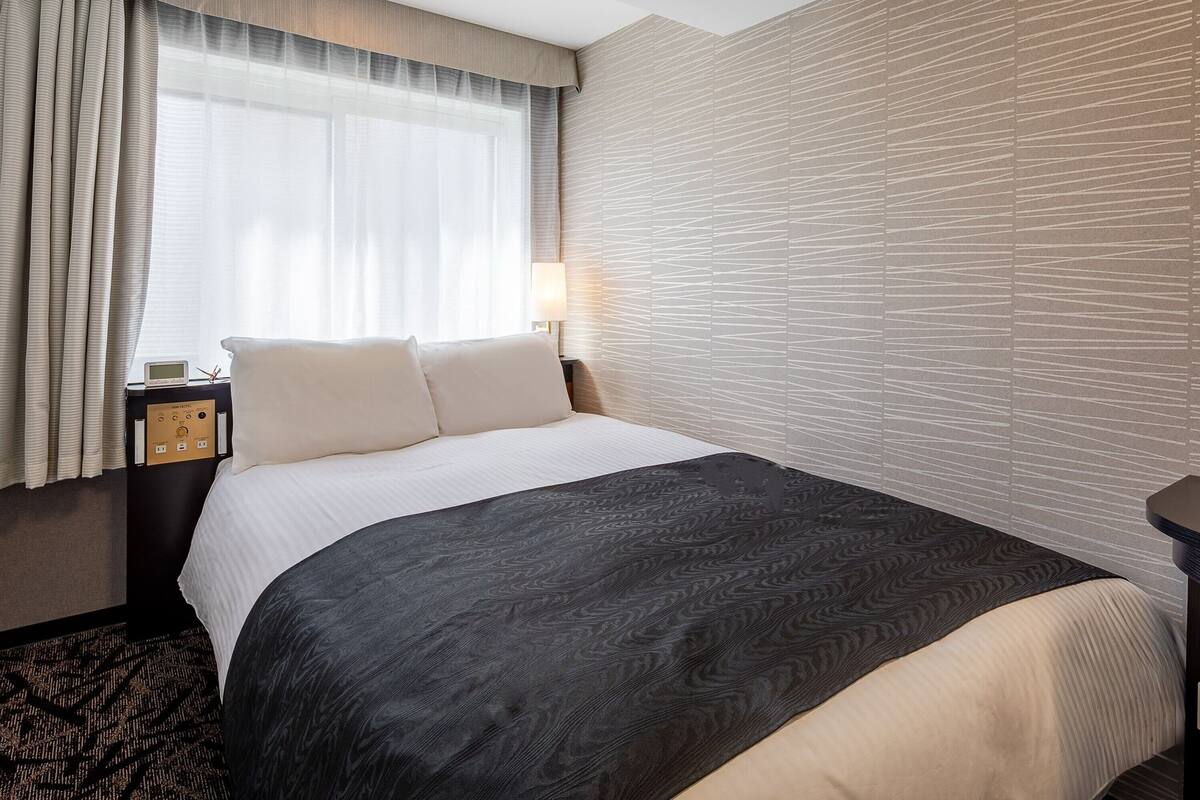 A hotel room with a bed, a nightstand with a clock, a lamp, and a window with curtains.