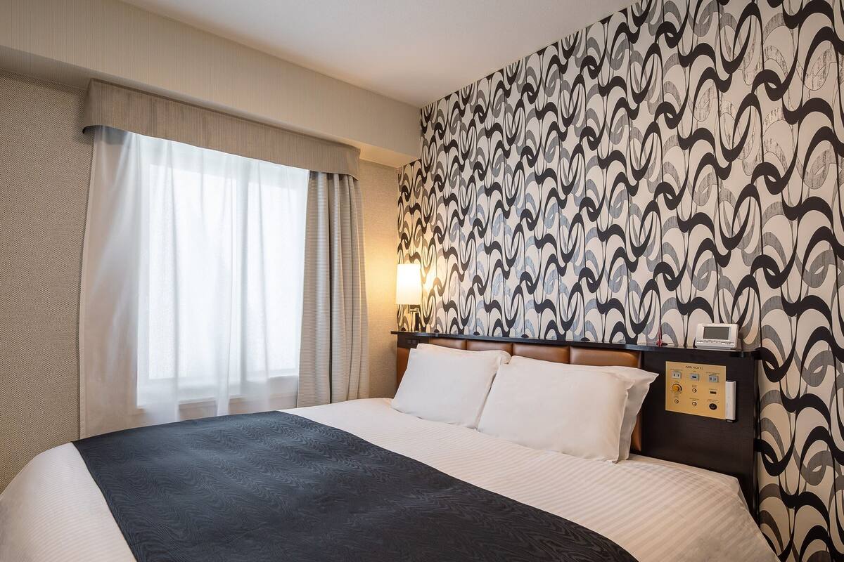 A hotel room with a bed, curtains, a patterned wall, a nightstand, and a lamp.