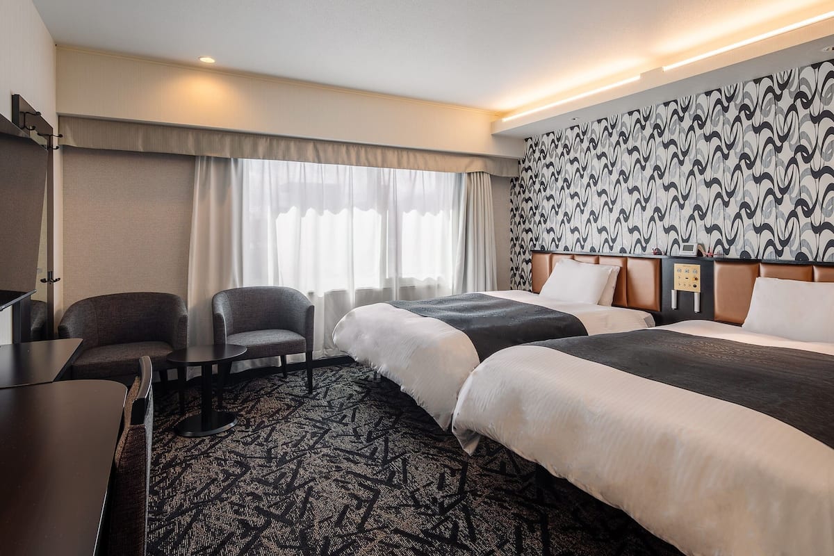 A hotel room with two beds, a desk, a chair, and a patterned carpet.