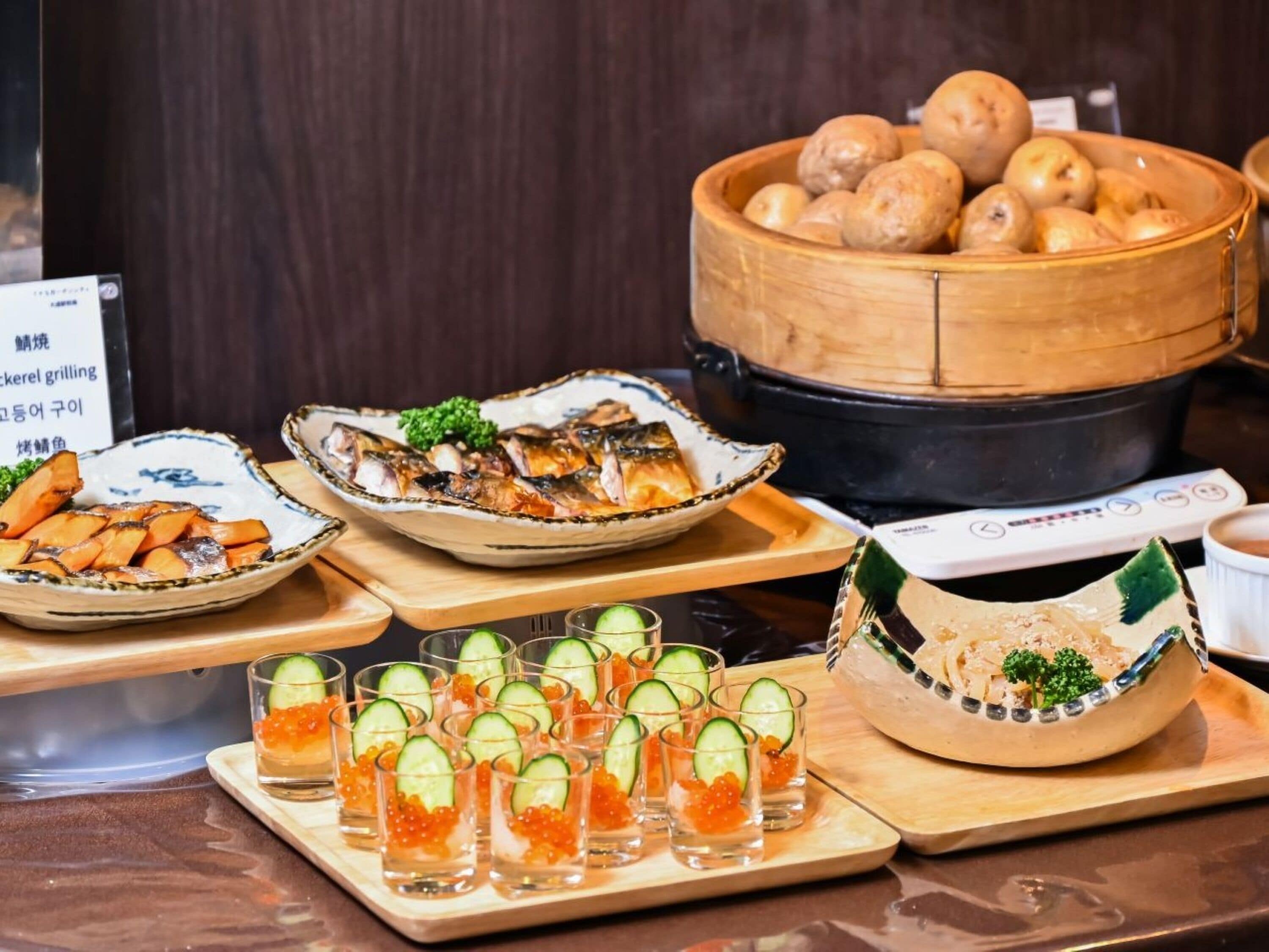 A buffet with various dishes, including grilled fish, potatoes, and a drink station with cucumber and salmon roe.