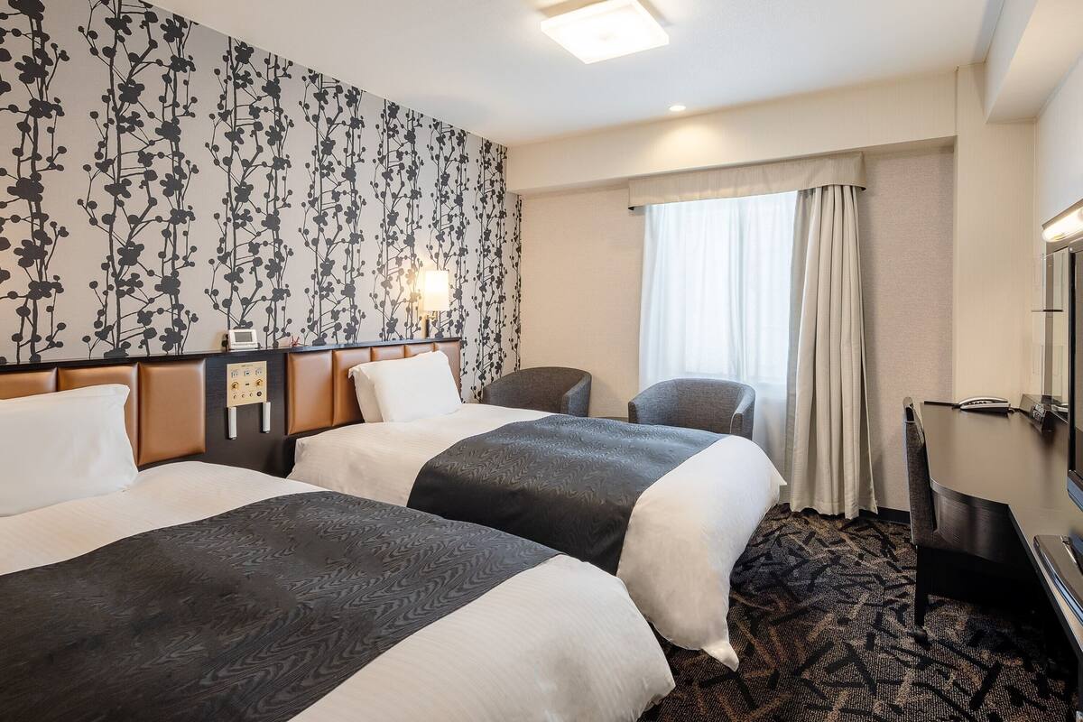 A hotel room with two beds, a desk, a chair, a window with curtains, and a wall with a floral pattern.