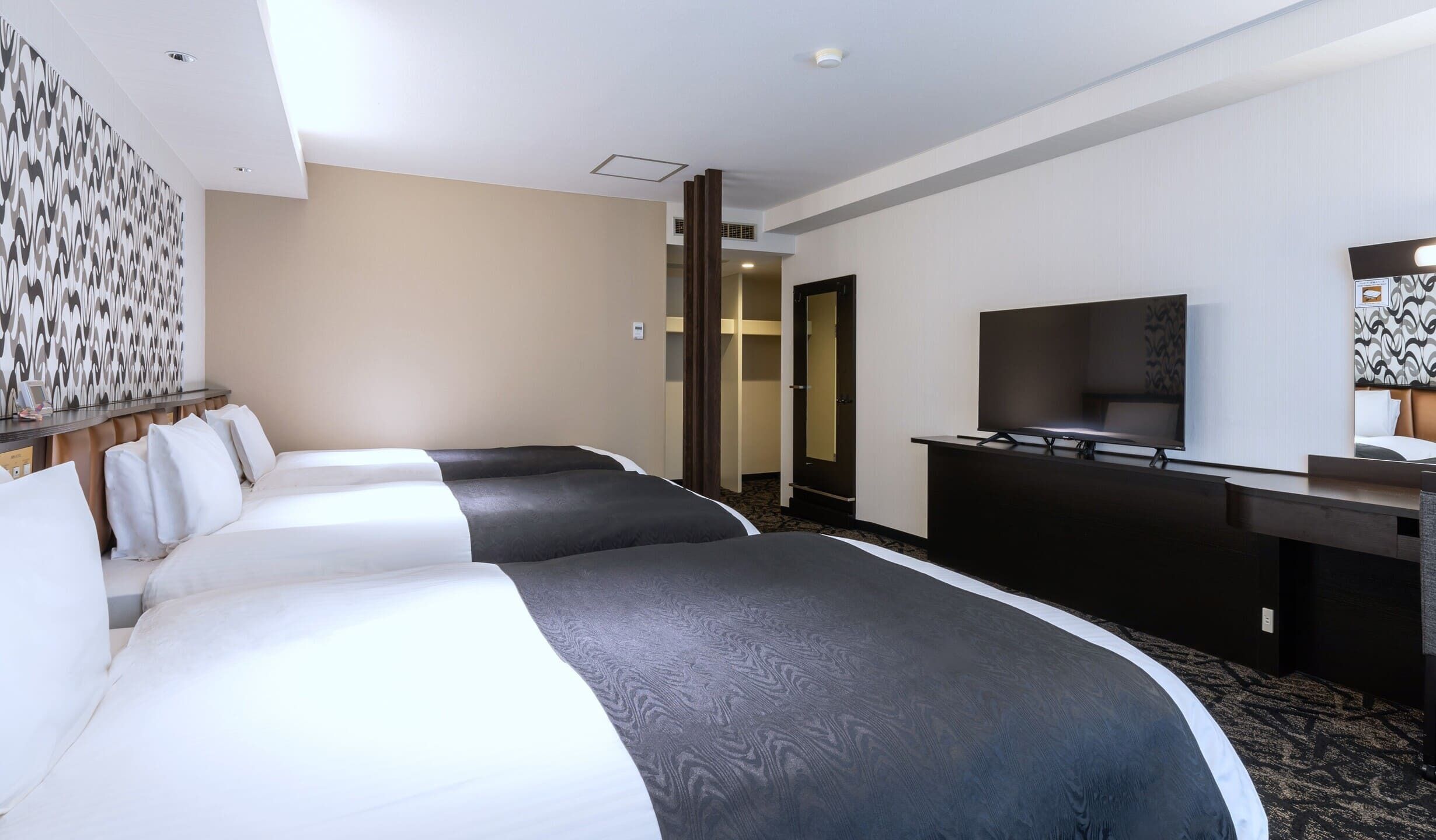 A hotel room with three beds, a TV, a desk, and a patterned wall.