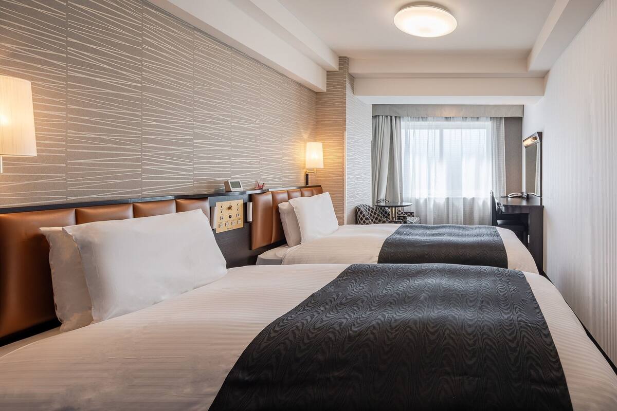 A hotel room with two beds, a desk, a chair, a lamp, and a mirror.