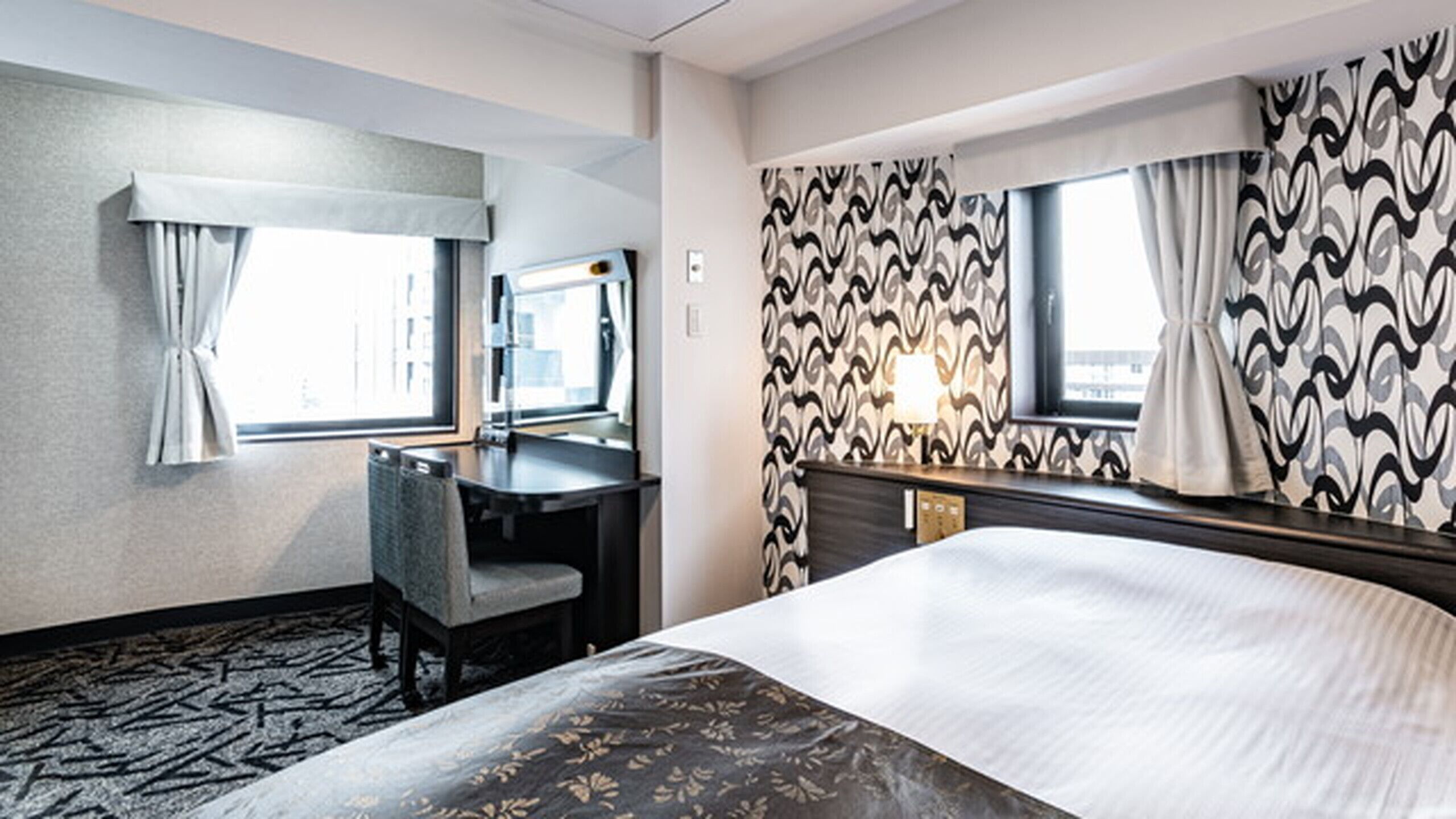 A hotel room with a bed, a desk, a chair, a window, and a patterned wall.