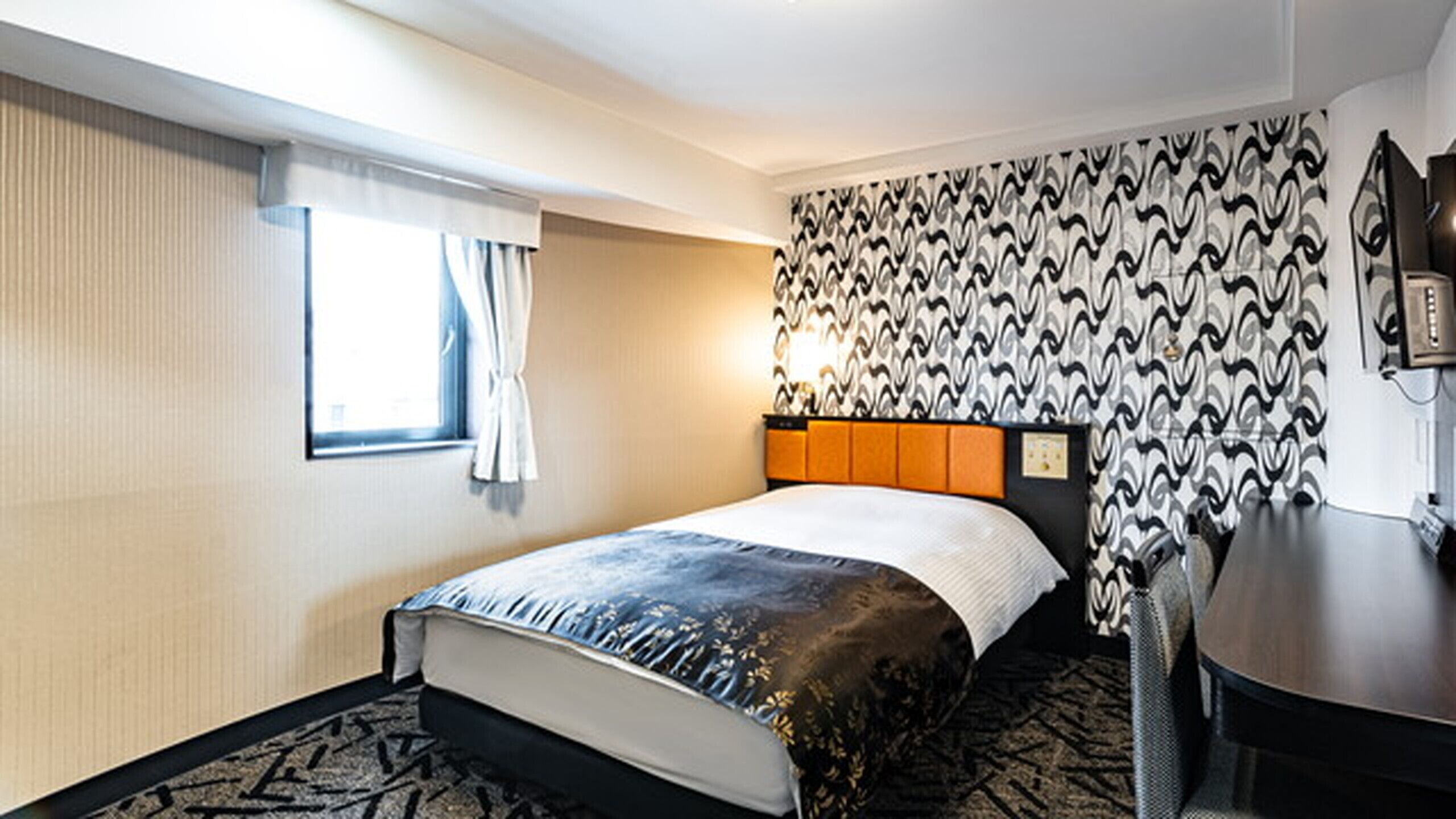 A hotel room with a bed, a desk, a chair, a window with curtains, and patterned wallpaper.