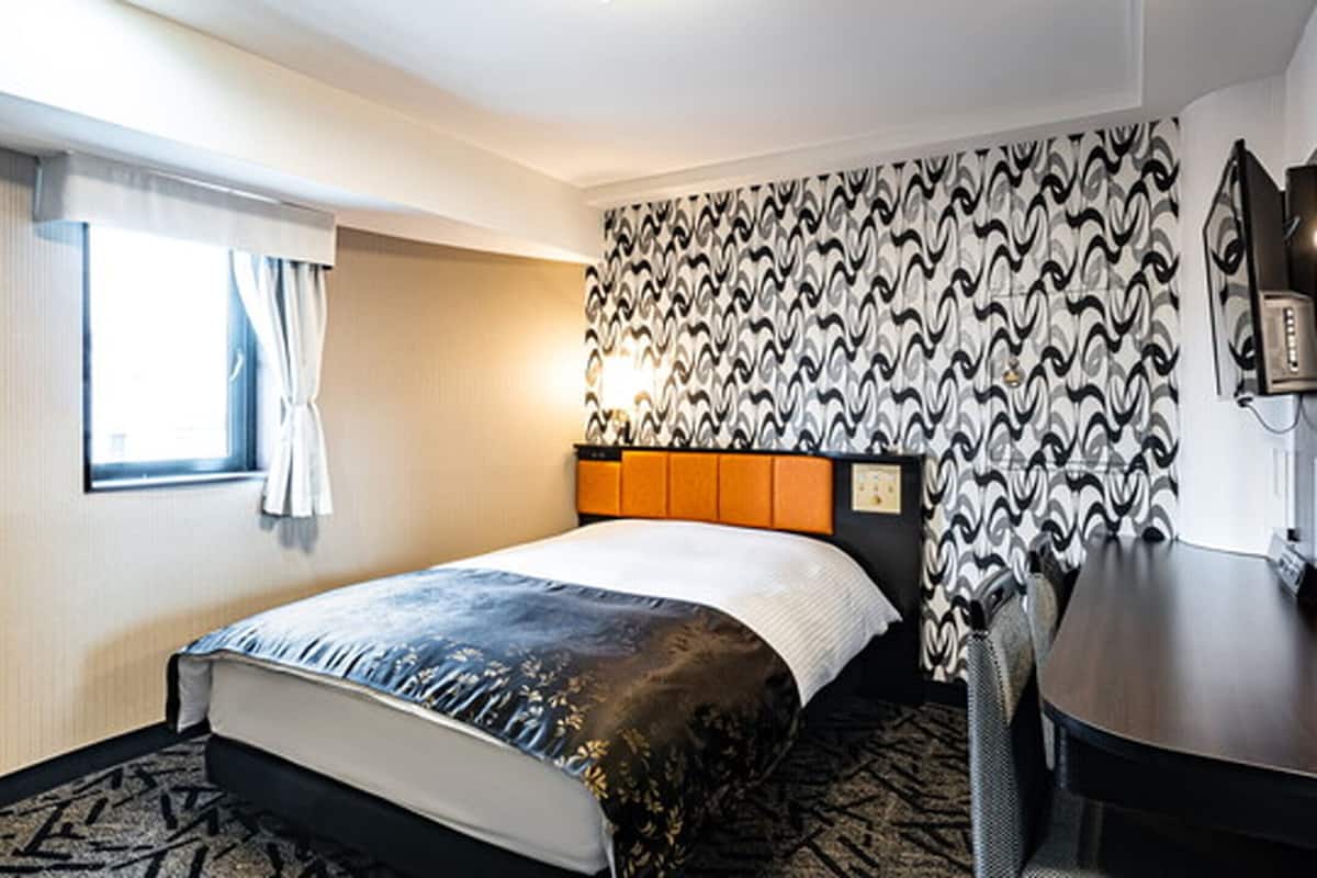 Ein Hotelzimmer mit Bett, Schreibtisch, Stuhl, einem Fenster mit Vorhängen und strukturierten Tapeten.