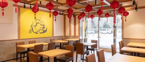 A restaurant interior with wooden tables and chairs, red lanterns hanging from the ceiling, and a yellow wall with a black illustration.