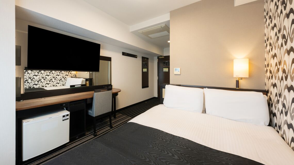 A hotel room with a bed, a desk, a chair, a television, and a wall-mounted lamp.