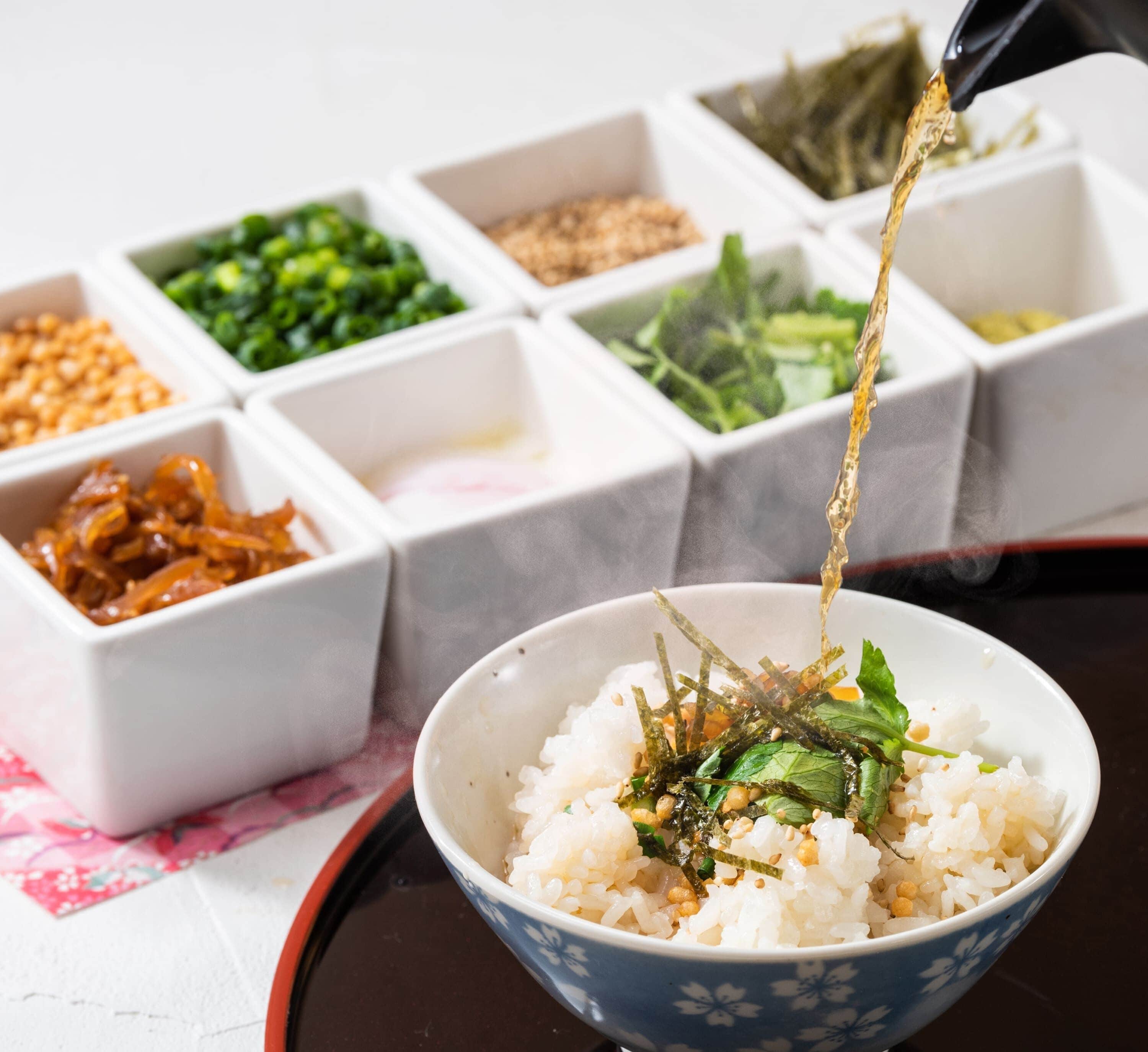 A bowl of rice with seaweed and greens, with various condiments in the background.