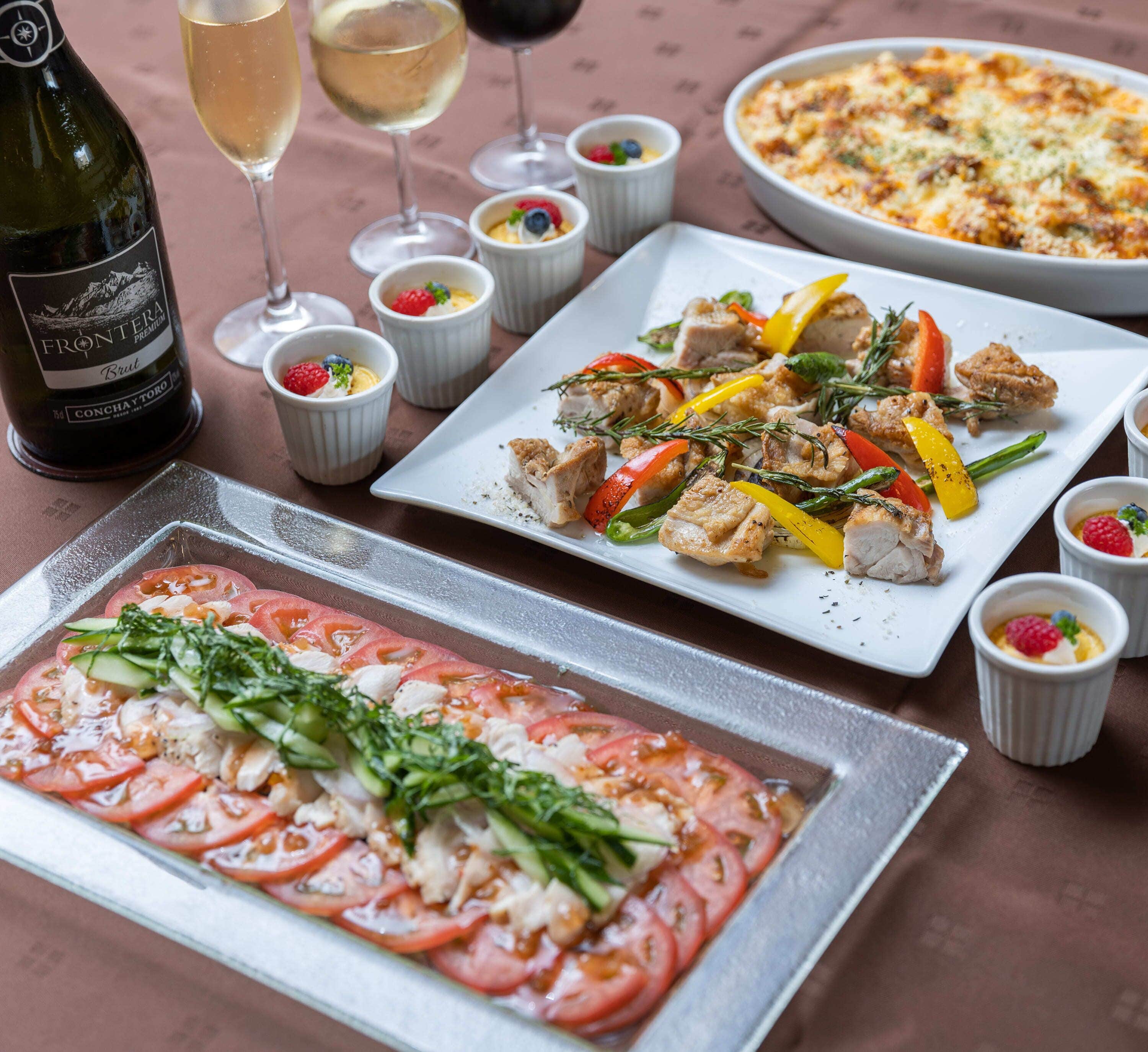 A dining table with various dishes, including a pizza, a plate of chicken with vegetables, and a tray of sliced tomatoes. There are also glasses of wine and small dessert cups.