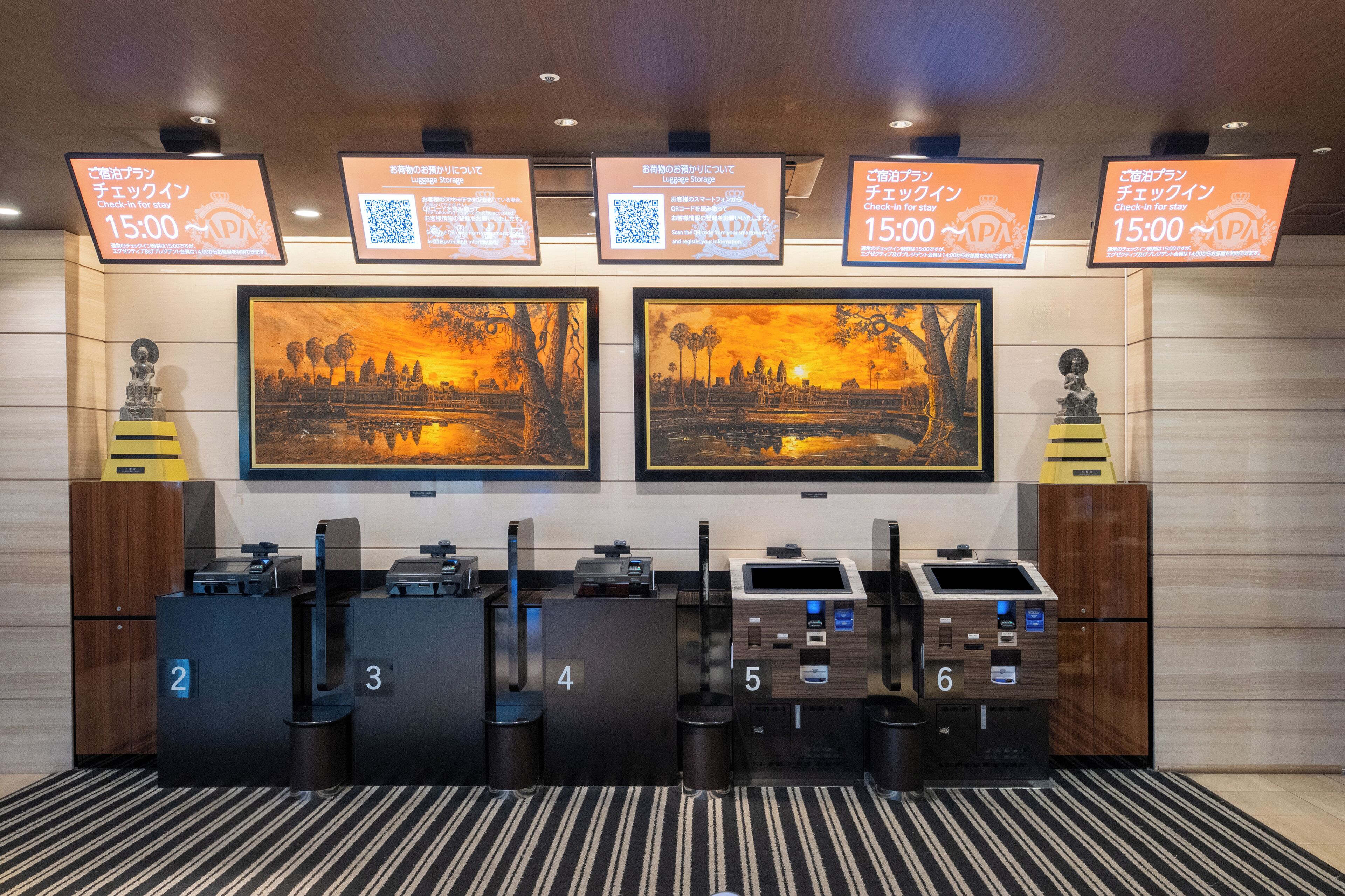 A hotel reception area with numbered desks, digital check-in displays, and artwork depicting a sunset scene.
