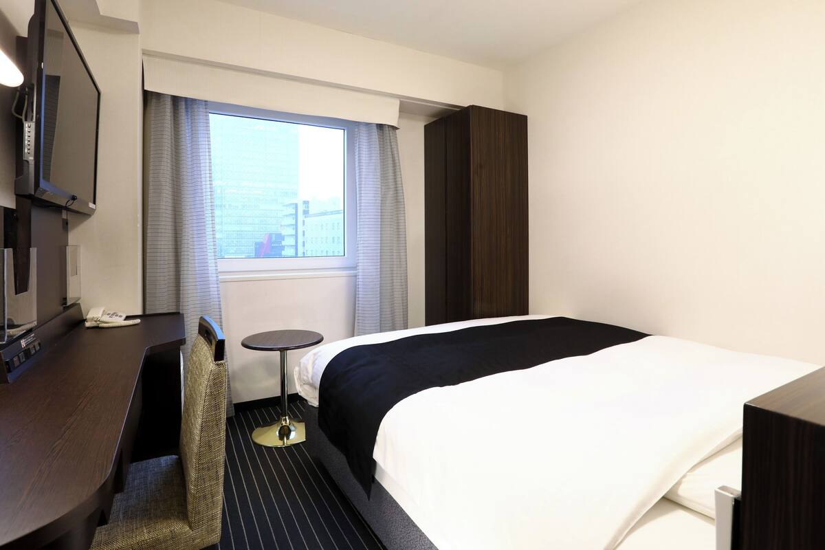 A hotel room with a bed, a desk with a telephone, a chair, a small table, and a TV mounted on the wall.