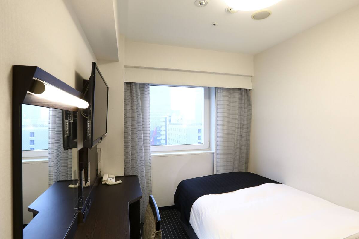 A hotel room with a bed, a desk with a telephone, a mirror, and a window with curtains.