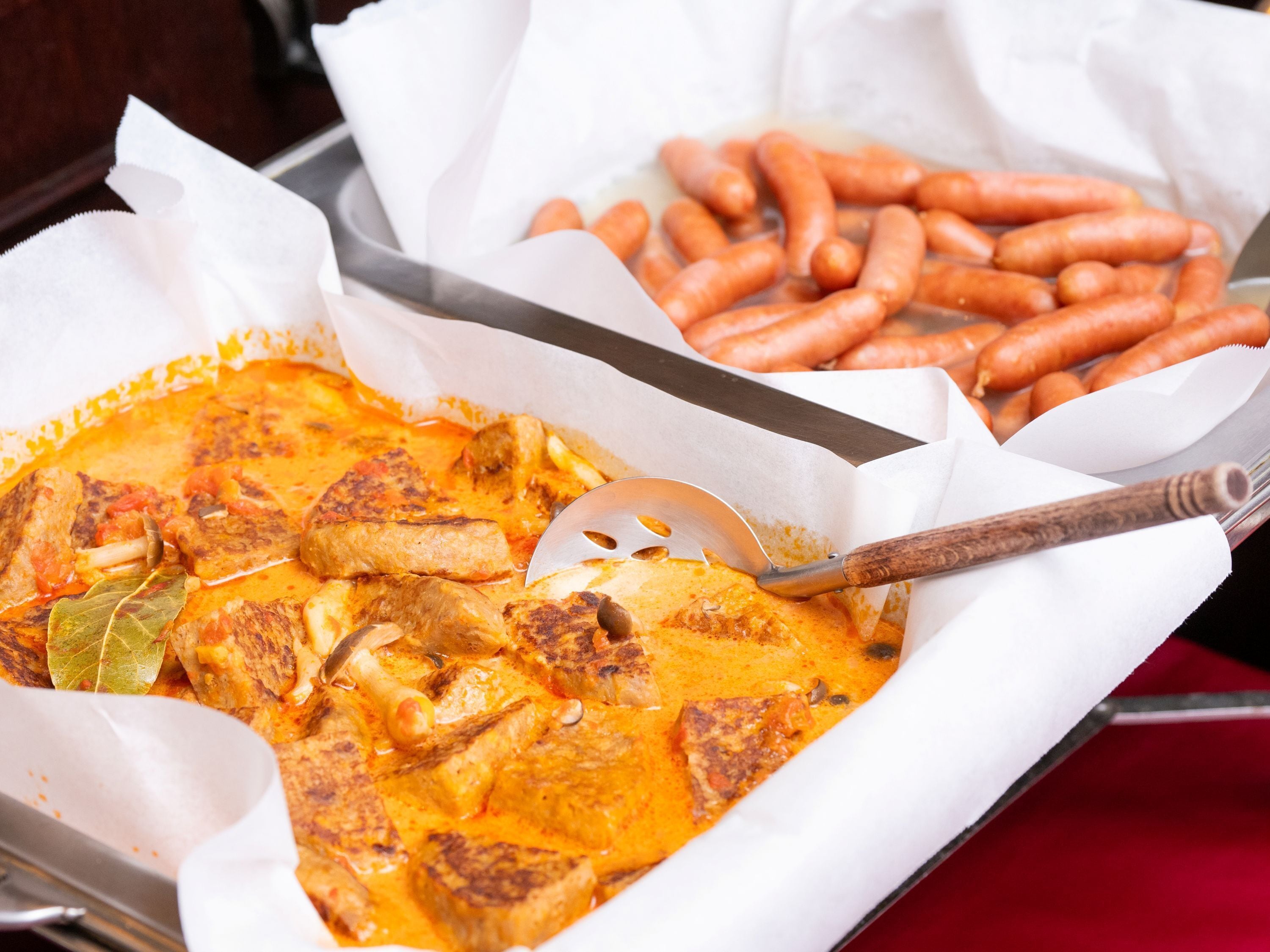 A tray with a curry dish, a wooden spatula, and a basket of sausages.