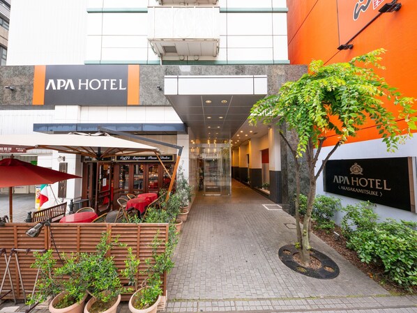 The exterior of APA Hotel with a covered outdoor seating area, potted plants, and a clear pathway leading to the entrance.