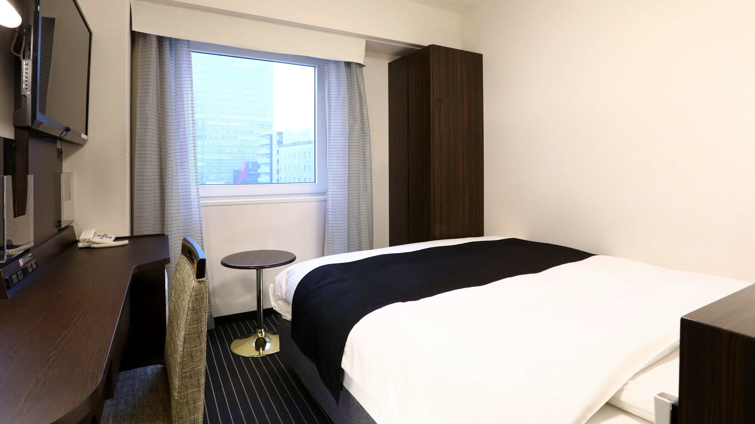 A hotel room with a bed, a small table, a chair, a desk with a phone, and a window with curtains.