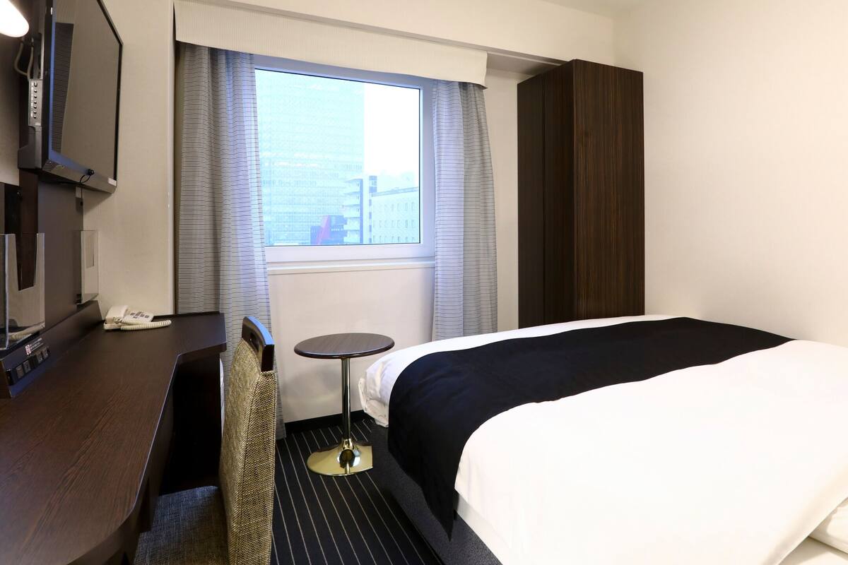A hotel room with a bed, a small table, a chair, a desk with a phone, and a window with curtains.